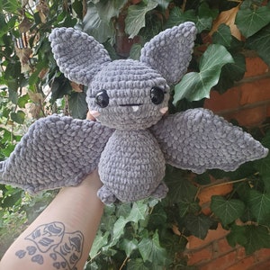 Puede incluir: Un juguete de peluche de murciélago de crochet gris con ojos negros y mejillas rosadas. El murciélago tiene orejas grandes y caídas y una gran envergadura.