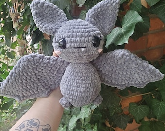 Puede incluir: Un juguete de peluche de murciélago de crochet gris con ojos negros y mejillas rosadas. El murciélago tiene orejas grandes y caídas y una gran envergadura.