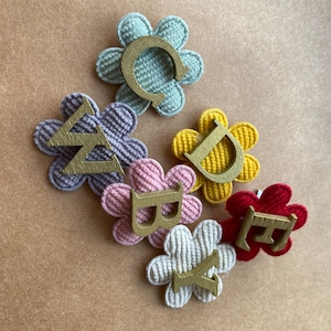 May include: Six flower-shaped fabric patches with letters in the centre. The letters spell out the word "baby". The flowers are in various colours, including pink, red, yellow, white, and blue.