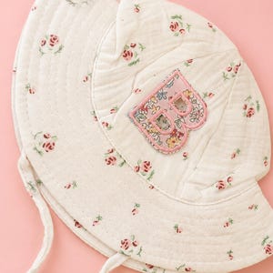 May include: White floral print sun hat with a pink letter "B" patch. The hat has a wide brim and a chin strap. The hat is decorated with small pink roses and green leaves.
