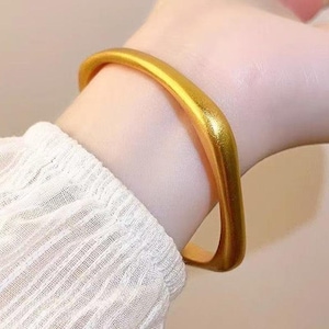 May include: A gold-colored bracelet with a rounded, rectangular shape. The bracelet is worn on a wrist, and the image shows a close-up view of the jewelry. The bracelet appears to be made of a solid metal and has a smooth, polished surface.