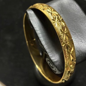 May include: A gold-colored bangle bracelet with a repeating starburst pattern. The bracelet is hinged and appears to be made of metal. The surface has a textured finish, and the bracelet is displayed on a dark surface.
