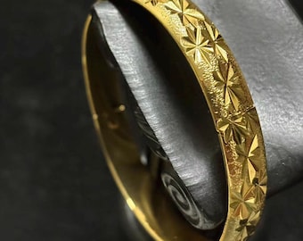 Peut inclure: Un bracelet jonc doré avec un motif d'éclats d'étoiles répété. Le bracelet à charnière semble être en métal. La surface a une finition texturée, et le bracelet est présenté sur une surface sombre.