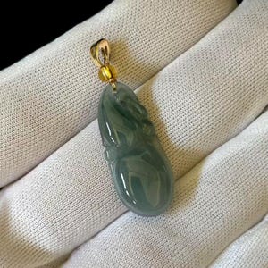 May include: A smooth, translucent, sage green jade pendant carved into an elongated, rounded shape. The pendant features a polished surface and is attached to a small, circular gold-toned metal bail at the top for hanging on a chain.
