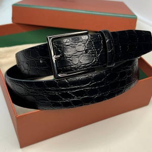 May include: A black belt with a textured, crocodile-like pattern. The belt features a silver-colored, square buckle and is presented in an orange box with a green and brown lid.