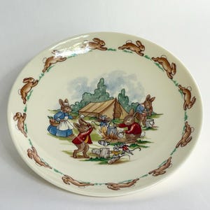 Könnte beinhalten: Eine cremefarbene Keramikplatte mit einer bezaubernden Illustration von Kaninchen beim Camping. Die zentrale Szene zeigt Kaninchen um ein Lagerfeuer, mit einem Zelt und Bäumen im Hintergrund. Ein Rand aus laufenden Kaninchen umkreist die Platte.