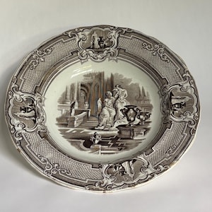 May include: A round, antique plate with a detailed brown and white design. The center features a classical scene with figures and architecture. The rim has an ornate border. A decorative piece for display.