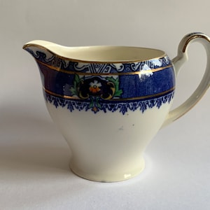 May include: A white ceramic milk jug with a blue and gold floral design. The jug has a rounded body and a small spout.