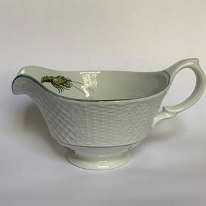 May include: White porcelain gravy boat with a woven basket pattern. The rim is trimmed with green, and a green and brown lobster illustration is on the inside. Features a handle and a footed base.