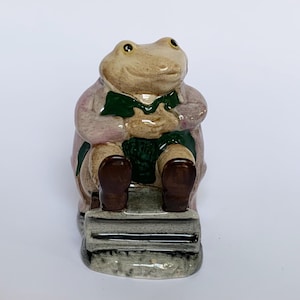 May include: A ceramic frog figurine wearing a pink coat and green waistcoat, seated on a small, grey, stone-like structure. The frog has brown boots and a neutral expression. The figurine is set against a white background.