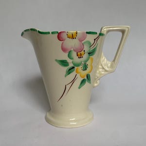 Op de afbeelding: Crèmekleurige keramische kan met een uitlopende vorm en een handvat. De kan is versierd met roze, gele en groene bloemmotieven. De rand heeft een groene geschulpte rand. Een decoratief item voor in huis.