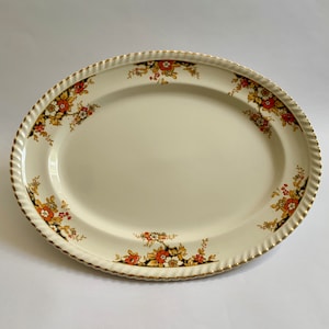 Pode incluir: Uma travessa oval branca para servir com uma borda dourada e um desenho floral em tons de laranja, castanho e branco.