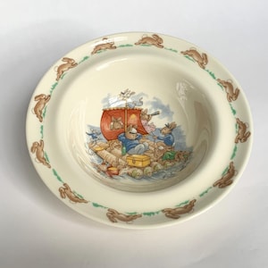 Könnte beinhalten: Cremefarbene Keramikschale mit einem Rand, der mit braunen Kaninchenillustrationen und einem grünen Rand verziert ist. Das Innere der Schale zeigt eine skurrile Szene von Kaninchen auf einem Floß mit rotem Segel, das auf dem Wasser fährt.