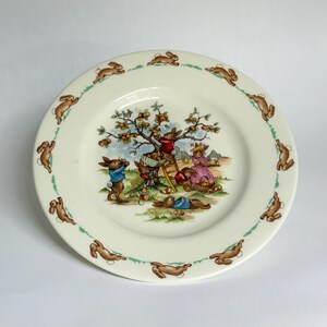 Könnte beinhalten: Eine cremefarbene Keramikplatte mit einer detaillierten Illustration von Hasen, die Früchte unter einem Baum sammeln. Der Rand der Platte zeigt ein sich wiederholendes Muster aus braunen Kaninchensilhouetten auf einer grünen gepunkteten Linie. Die Szene spielt in einer ländlichen Landschaft.