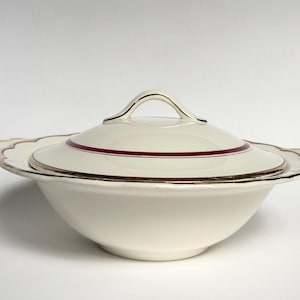 May include: Cream-coloured lidded serving bowl with a scalloped rim and gold trim. A thin burgundy stripe accents the lid and the rim. The handle on the lid is arched and gold-toned. A classic piece for serving.
