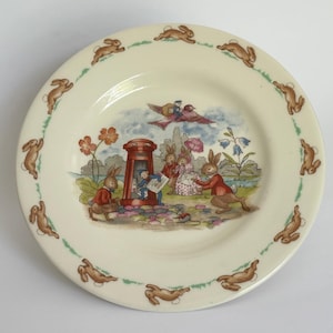 Könnte beinhalten: Cremefarbene Keramikplatte mit einer skurrilen Illustration von Kaninchen in einer Gartenszene. Der Teller hat einen Rand mit laufenden Kaninchen und ein zentrales Bild mit einem roten Briefkasten und floralen Elementen.