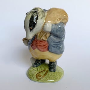 May include: A ceramic figurine of a badger dressed in a waistcoat, jacket, and carrying a sack. The badger is standing on a green base. The figurine has a detailed, hand-painted appearance with a variety of colors.