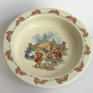 Könnte beinhalten: Cremefarbene Keramikschale mit einem Rand, der mit braunen Kaninchenillustrationen verziert ist. Die Mitte zeigt eine Szene von Kaninchen, die in der Nähe eines Zeltes campen. Die Kaninchen sind bekleidet und versammeln sich um ein Lagerfeuer. Ein charmantes, Vintage-Design.