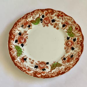 May include: A white ceramic plate with a brown, orange and green floral pattern around the rim.