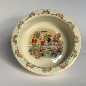 Könnte beinhalten: Cremefarbene Keramikschale mit einem Rand, der mit braunen Kaninchenillustrationen verziert ist. Das Innere der Schale zeigt eine Szene von Kaninchen in Kleidung, die Holz und andere Gegenstände tragen. Ein charmantes, skurriles Design.