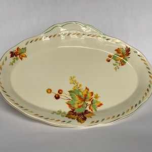 May include: Cream-coloured oval serving platter with a scalloped edge and autumn leaf design. The rim features a repeating pattern of gold, green, and orange. The platter has a handle on one side and is decorated with clusters of leaves and berries.