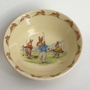 Könnte beinhalten: Eine cremefarbene Keramikschale mit einer Illustration von Hasen beim Golfspielen. Der Rand der Schale zeigt ein sich wiederholendes Muster von laufenden Hasen. Im Inneren spielen drei Hasen Golf auf einem grünen Feld.