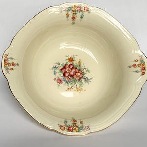 May include: Cream-coloured vintage serving bowl with a floral design. The bowl has a scalloped edge and gold trim. The floral pattern features red, pink, and yellow flowers with green leaves. The bowl has a square shape with rounded corners.