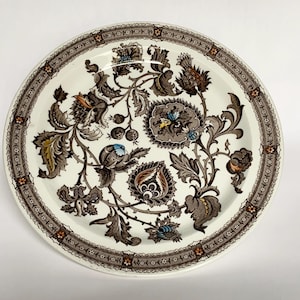 Może przedstawiać: Okrągły, biały ceramiczny talerz z szczegółowym wzorem kwiatowym w kolorach brązu, beżu i niebieskiego. Obręcz talerza ma dekoracyjną obwódkę. Wzór zawiera stylizowane kwiaty, liście i inne elementy botaniczne.