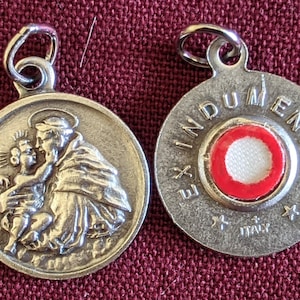 Peut inclure: Pendentif en argent représentant Saint Antoine de Padoue tenant l'Enfant Jésus. Au dos du pendentif se trouve un reliquaire rouge et blanc avec le texte "Ex Indumentis" et "Italie".