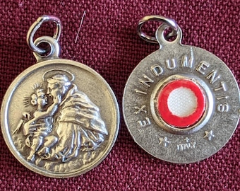 May include: Silver pendant with a depiction of Saint Anthony of Padua holding the Christ Child. The back of the pendant has a red and white relic holder with the text "Ex Indumentis" and "Italy".