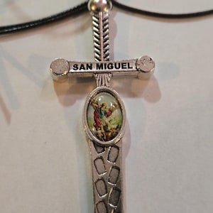 May include: A silver pendant necklace with a sword design. The crossguard is inscribed with the words "SAN MIGUEL". The pendant has an oval centre with a colourful image. The necklace is strung on a black cord.