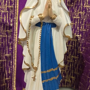 Könnte beinhalten: Statue einer Frau in einem weißen Gewand mit goldenen Verzierungen, einem blauen Schärpe und einem goldenen Rosenkranz. Die Statue hat einen ruhigen Ausdruck und gefaltete Hände. Der Hintergrund ist ein violetter Vorhang.