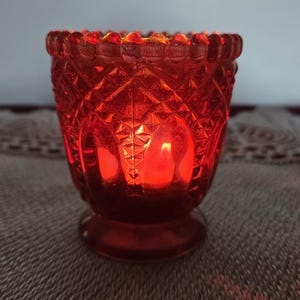 Pode incluir: Um porta-velas de vidro vermelho com um padrão de diamante. O porta-velas tem uma base larga e uma parte superior estreita. A vela está acesa e brilhando.