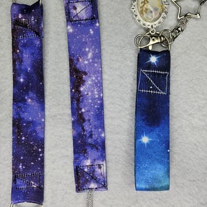 May include: Three fabric straps featuring a deep blue and purple galaxy print. The left strap has a silver ring and star charms. The center strap includes a purple plastic clip and a grey cord. The right strap features a star charm and an oval pendant.