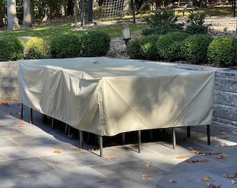 Puede incluir: Una mesa rectangular de exterior cubierta con una funda protectora beige. La mesa tiene patas de metal negro y está situada en un patio de piedra. El fondo presenta arbustos y árboles verdes.