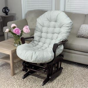 May include: A light gray upholstered glider chair with a dark brown wooden frame. The chair has a tufted back and seat cushion. The chair is in a living room setting with a sofa and a small table with flowers.