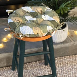 May include: A round seat cushion with a botanical pattern in shades of green, white, and tan, tied to a green wooden stool with an orange seat. The cushion has a tufted design and is placed outdoors.