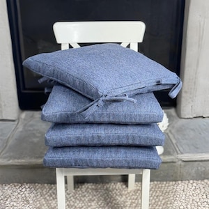 May include: Four square blue seat cushions stacked on a white chair. The cushions have a textured appearance and are tied with fabric ties. The chair is positioned in front of a dark background.