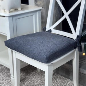 May include: A square, dark gray, textured fabric chair cushion with fabric ties at the back corners, secured onto a white wooden chair with an X-shaped backrest. The cushion has a woven appearance and sits flat on the seat of the chair.