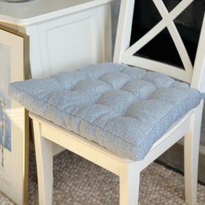 May include: A square, light blue tufted seat cushion with a textured fabric finish. The cushion features a grid of stitched indentations across its surface, creating a padded, raised appearance. It is placed flat on a white wooden chair.
