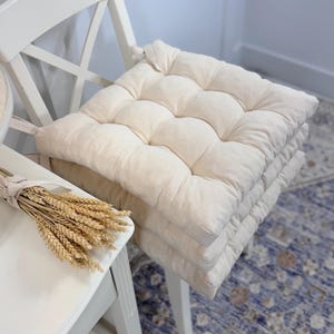May include: A stack of four cream-colored square seat cushions with a tufted design. The cushions are on a white chair. A bundle of wheat tied with a ribbon sits on a white table next to the chair.