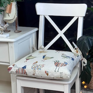 May include: A square cushion with a white background and a colorful bird pattern rests on a white chair. The birds are depicted in various styles and colors, including blue, red, and brown. The cushion is tied to the chair with fabric ties.