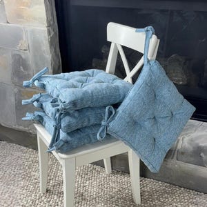 May include: A stack of light blue square seat cushions with tie closures, resting on a white wooden chair. The cushions have a textured appearance and are arranged near a stone fireplace.