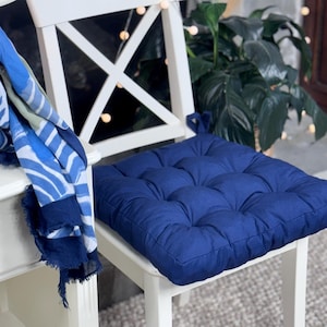 May include: A navy blue tufted cushion sits on a white chair. A blue and white patterned scarf hangs on the side. The cushion is square and has a textured, quilted appearance. The chair has an X-shaped back.