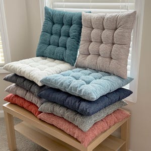 May include: A stack of tufted seat cushions in various colors, including teal, beige, white, light blue, navy, gray, and coral. The cushions are arranged on a light-colored wooden bench near a window with white blinds. The cushions have a square, button-like design.