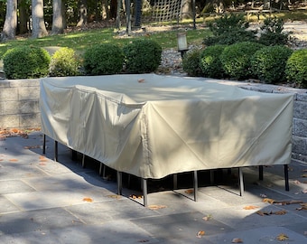 Puede incluir: Una funda beige para muebles de patio está colocada sobre una mesa y sillas. La funda está hecha de un material resistente al agua y tiene un cierre de cordón. La funda está sobre un patio gris.