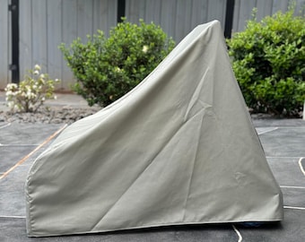 Puede incluir: Una funda de silla de exterior de color gris claro. La funda está hecha de una tela duradera y resistente al agua y está diseñada para proteger la silla de los elementos. La funda está sobre un patio de piedra gris oscuro, con arbustos verdes y una valla de madera en el fondo.