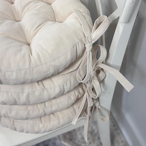 May include: A stack of four round, cream-colored chair cushions with tufted detailing. Each cushion has a fabric tie attached. The cushions are resting on a white chair.