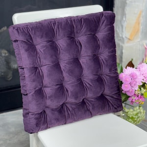 May include: A square, deep purple velvet seat cushion featuring a tufted design with multiple indentations across its surface. The cushion has a soft, plush texture and is positioned upright on a white chair, showcasing its thick, padded construction.