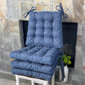 May include: Set of three blue square seat cushions with tufted detailing and attached fabric ties. The cushions are stacked on a white chair with a decorative back. The cushions are made of a textured fabric.